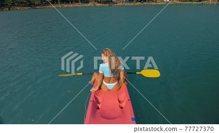 Seductive blonde woman in blue swimsuit sails on pink plastic kayak along azure sea at exotic resort backside upper above view. 77727370