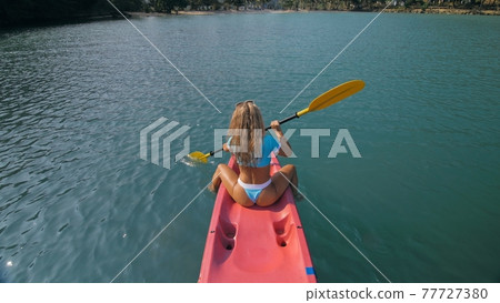 Seductive blonde woman in blue swimsuit sails on pink plastic kayak along azure sea at exotic resort backside upper above view. 77727380
