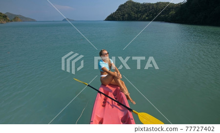 Pretty sportive blonde lady in stylish swimsuit sails on pink plastic canoe along azure ocean bay at exotic resort. 77727402