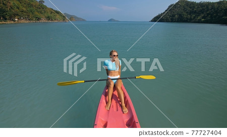 Pretty sportive blonde lady in stylish swimsuit sails on pink plastic canoe along azure ocean bay at exotic resort. 77727404