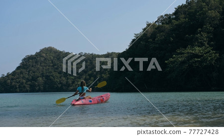 Young blonde woman in blue swimsuit rows pink plastic canoe along azure sea bay past island with palms under blue sky at resort. 77727428