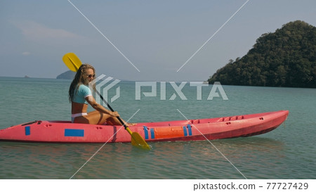 Young blonde woman in blue swimsuit rows pink plastic canoe along azure sea bay past island with palms under blue sky at resort. Young blonde woman in blue swimsuit rows pink plastic canoe along azure sea bay past island with palms under blue sky at resort. 77727429