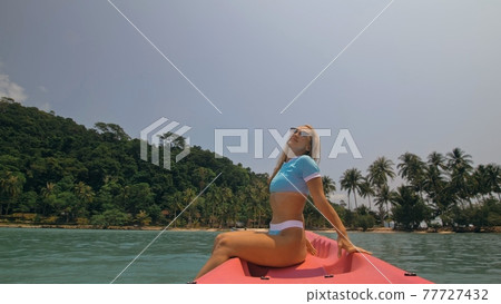 Pretty young blonde woman in stylish swimsuit and sunglasses sits on pink canoe sailing along sea against hills. 77727432