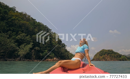 Pretty young blonde woman in stylish swimsuit and sunglasses sits on pink canoe sailing along sea against hills. 77727433