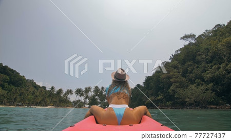 Sexy young woman in blue swimsuit and hat swings feet in water on pink canoe on sea against hills backside view. 77727437
