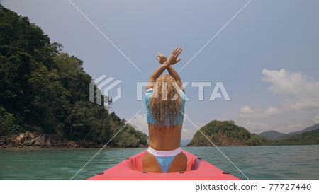Sexy young woman in blue swimsuit swings feet in water on pink canoe on sea against hills backside view. Sexy young woman in blue swimsuit swings feet in water on pink canoe on sea against hills backside view. 77727440