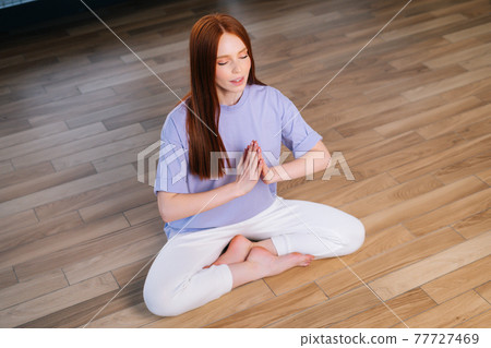 Young redhead woman meditating practicing breathing yoga exercise, sitting on floor near window in lotus position. 77727469