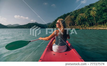 Long haired blonde woman with sunglasses rows bright pink canoe along sea bay water to beach with growing palms. 77727574