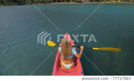 Seductive blonde woman in blue swimsuit sails on pink plastic kayak along azure sea at exotic resort backside upper above view. Seductive blonde woman in blue swimsuit sails on pink plastic kayak along azure sea at exotic resort backside upper above view. 77727883