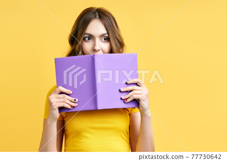 Young woman hiding behind an open book and looking to camera isolated over yellow background 77730642