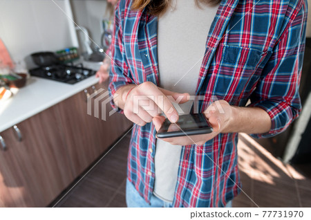 Close-up of the hand and body of a young guy in a plaid shirt at home holding a smartphone. He touches the screen with his thumb. Young man in the kitchen using his phone Close-up of the hand and body of a young guy in a plaid shirt at home holding a smartphone. He touches the screen with his thumb. Young man in the kitchen using his phone 77731970