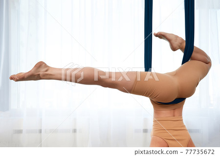 Cheerful sporty woman wearing top and leggings performing anti-gravity yoga exercise at spacious health club 77735672