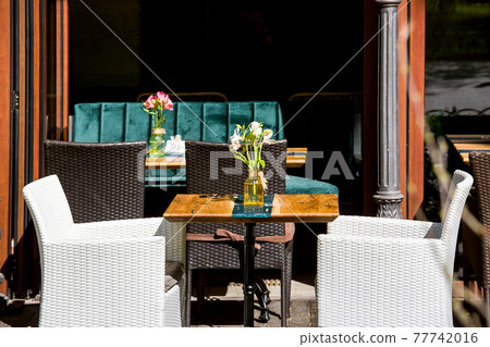 outdoor cafe terrace with wooden tables with a glass vase and a flower and wicker chairs in white and brown outside the restaurant with empty seats on summer sunny day, nobody. 77742016