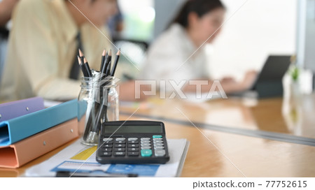 A calculator is putting on the wooden table surrounded by various office equipment over employees as a background. 77752615