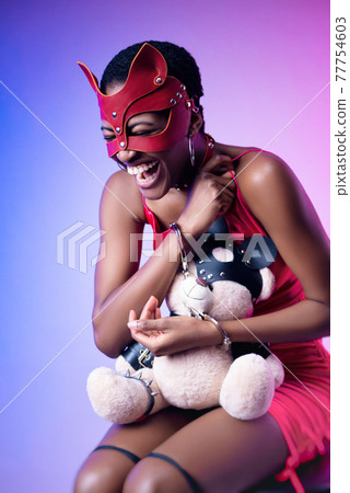 black woman in a bdsm cat mask laughs handcuffed with a teddy bear in her hands black woman in a bdsm cat mask laughs handcuffed with a teddy bear in her hands 77754603