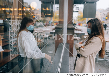 Two women in protective masks opposite each other, window between them. Two women in protective masks opposite each other, window between them. 77754944