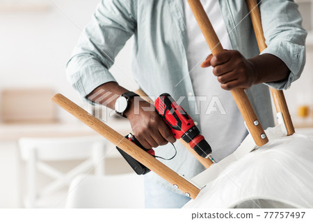 Furniture assembly process. African american handyman screwing legs to chair with electric screwdriver, crop 77757497