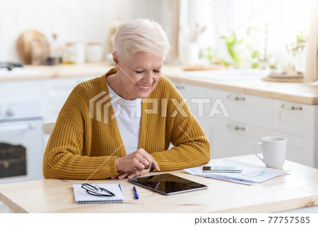 Joyful grey-haired senior woman using digital tablet in kitchen 77757585