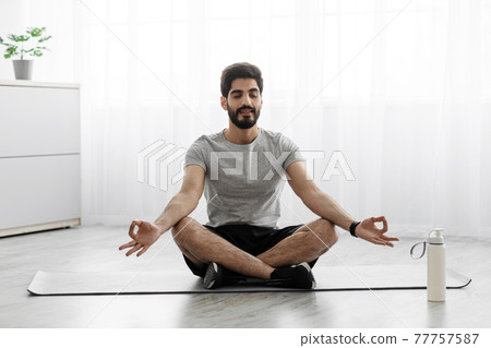 Cheerful arabian bearded millennial man sits on mat in lotus position, rest on floor with bottle of water Cheerful arabian bearded millennial man sits on mat in lotus position, rest on floor with bottle of water 77757587