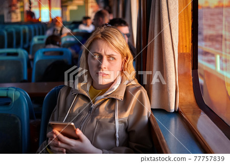 Passenger cabin of ferry, offended young woman sits by window with smartphone in her hands. 77757839