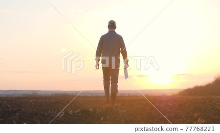 a farmer walks with a tablet in his hand on the ground at sunset in the sky, a working man agronomist is engaged in land cultivation in the field, it is time to fertilize the land for seedlings of the a farmer walks with a tablet in his hand on the ground at sunset in the sky, a working man agronomist is engaged in land cultivation in the field, it is time to fertilize the land for seedlings of the 77768221