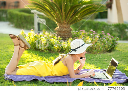 Girl student in yellow summer dress resting on green lawn in summer park studying on computer laptop having conversation on mobile cell phone. Doing business and learning during quarantine concept. Girl student in yellow summer dress resting on green lawn in summer park studying on computer laptop having conversation on mobile cell phone. Doing business and learning during quarantine concept. 77769603