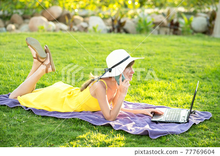 Girl student in yellow summer dress resting on green lawn in summer park studying on computer laptop having conversation on mobile cell phone. Doing business and learning during quarantine concept. Girl student in yellow summer dress resting on green lawn in summer park studying on computer laptop having conversation on mobile cell phone. Doing business and learning during quarantine concept. 77769604