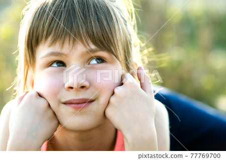 Portrait of pretty child girl with gray eyes and long fair hair leaning on her hands smiling happily outdoors on blurred bright background. Cute female kid on warm summer day outside. 77769700