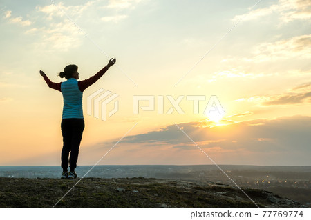 Silhouette of a woman hiker standing alone raising hands enjoying sunset outdoors. Female tourist on rural field in evening nature. Tourism, traveling and healthy lifestyle concept. Silhouette of a woman hiker standing alone raising hands enjoying sunset outdoors. Female tourist on rural field in evening nature. Tourism, traveling and healthy lifestyle concept. 77769774