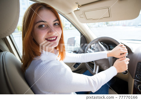 Young redhead woman driver behind a wheel driving a car smiling happily. 77770166
