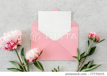 Mockup invitation, blank paper greeting card, pink envelope and peonies on gray stone table. Flower background. Flat lay, top view. 77770435