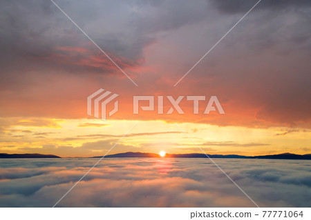 Aerial view of yellow sunset over white puffy clouds with distant mountains on horizon. 77771064