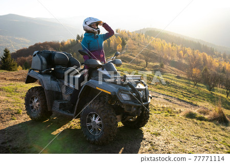 Young happy female driver in protective helmet enjoying extreme ride on ATV quad motorbike in fall mountains at sunset. 77771114