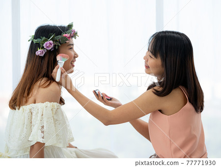 Two Asian teenagers making up each other cheek by brush on bed in bright bedroom 77771930