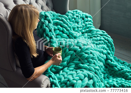 A woman in a black jumper holds a cup of tea and looks out the window, covered by a green merino blanket. A woman in a black jumper holds a cup of tea and looks out the window, covered by a green merino blanket. 77773416