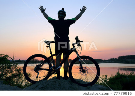 silhouette of a standing cyclist with arms raised with a bike at sunset. silhouette of a standing cyclist with arms raised with a bike at sunset. 77779916