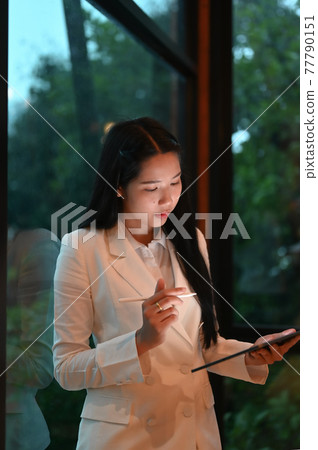 A businesswoman is holding a computer tablet and stylus pen while standing over the office glass wall. A businesswoman is holding a computer tablet and stylus pen while standing over the office glass wall. 77790151