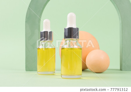 Mockup image of dropper bottles with polyglutamic, hyaluronic acid, serum, moisturizer, facial anti-aging oil. Trendy still life composition with modern shapes against green background with copy space 77791312