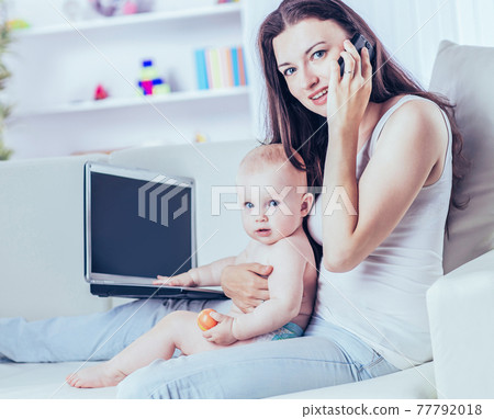 young mother holding a year-old baby in his arms,talking on his cell phone and working on laptop young mother holding a year-old baby in his arms,talking on his cell phone and working on laptop 77792018