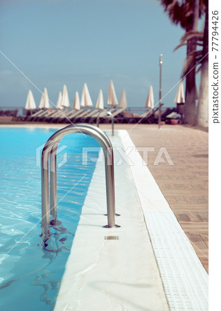 Hotel swimming pool side with blue water, close-up shot, with sunbeds on background Hotel swimming pool side with blue water, close-up shot, with sunbeds on background 77794426