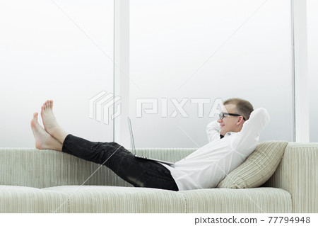 modern man with laptop lying on sofa in living room modern man with laptop lying on sofa in living room 77794948