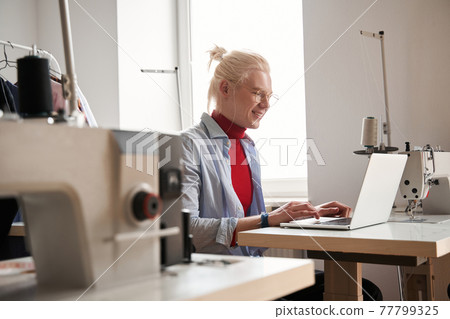 Man looking at the laptop screen while working at the table near sewing machine Man looking at the laptop screen while working at the table near sewing machine 77799325
