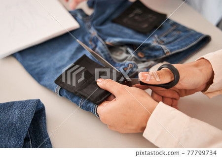 Woman hands cutting fabric with scissors at the table with jeans at the background Woman hands cutting fabric with scissors at the table with jeans at the background 77799734