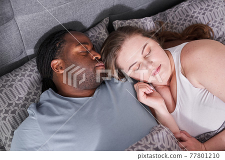 Multiracial couple laying at the bed indoors at home and hugging while sleeping 77801210