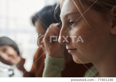 Girl brushing her eyelashes with mascara while her grandmother looking at the mirror Girl brushing her eyelashes with mascara while her grandmother looking at the mirror 77801286