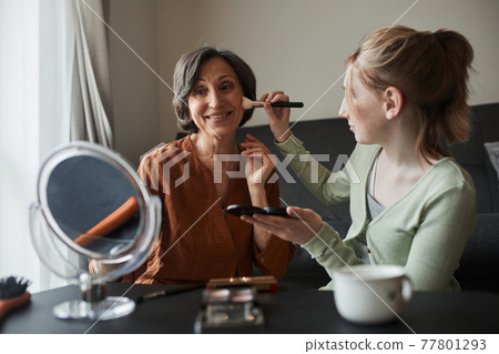 Woman looking at the mirror with pleasure while her granddaughter preparing makeup for her Woman looking at the mirror with pleasure while her granddaughter preparing makeup for her 77801293