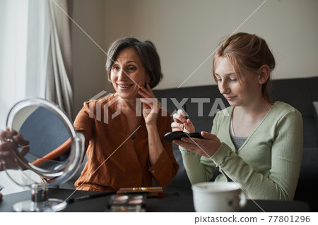 Woman looking at the mirror while her granddaughter preparing makeup for her Woman looking at the mirror while her granddaughter preparing makeup for her 77801296