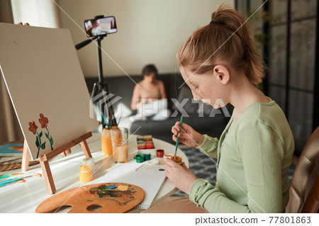 Girl artist sitting in front of the smartphone at the tripod in her living room Girl artist sitting in front of the smartphone at the tripod in her living room 77801863