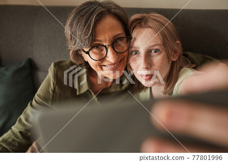 Woman making funny faces with her freckled granddaughter while taking selfie at home 77801996
