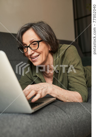 Grandmother wearing glasses lying at the sofa with laptop Grandmother wearing glasses lying at the sofa with laptop 77802040